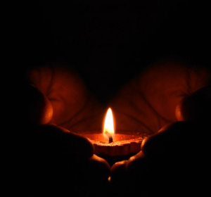 Na zdjęciu świeczka trzymana w dłoniach, której płomień rozświetla ciemność